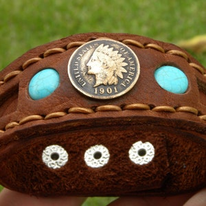May include: Brown leather bracelet with a gold-toned coin featuring an image of an American Indian head and the text "United States of America 1901". The bracelet also has two turquoise stones.