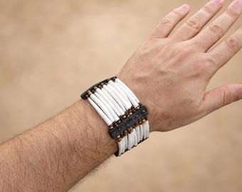 May include: A wide bracelet featuring multiple rows of white cylindrical beads arranged vertically, separated by dark brown leather spacers and accented with small, round, metallic-toned beads at the connection points.
