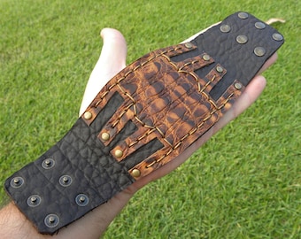 May include: A brown leather bracelet with a decorative design featuring a brown and black leather pattern. The bracelet has multiple brass studs and stitching.