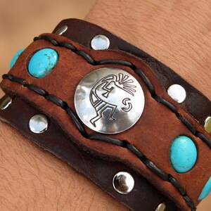 Pode incluir: Uma pulseira de couro castanho larga com um concho circular prateado ao centro, apresentando uma gravação de uma figura Kokopelli. A pulseira é decorada com pedras turquesas redondas, tachas metálicas prateadas e atacadores de couro preto.