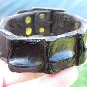 Puede incluir: Un brazalete de cuero negro con un diseño texturizado, similar al de un cocodrilo. El brazalete tiene un cierre de hebilla y está acentuado con herrajes dorados.