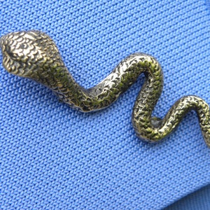 May include: A gold-coloured snake cufflink with a textured surface. The snake is curled in an S-shape.