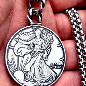 Pode incluir: Um pingente de moeda de prata com um relevo detalhado de uma mulher, o texto "IN GOD WE TRUST" e o ano de 2000. O pingente está preso a um colar de corrente de prata.