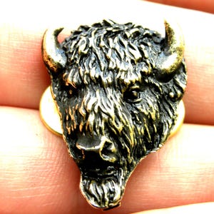 May include: A detailed, bronze-toned buffalo head pin. The pin features a textured, fur-like appearance with prominent horns and a detailed facial structure. The pin is set on a gold-colored backing.