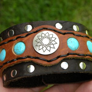 Puede incluir: Una pulsera ancha de cuero en marrón y negro. Presenta piedras de color turquesa, tachuelas plateadas y un emblema central plateado con un diseño de estrella. La pulsera mide aproximadamente 5 cm de ancho.