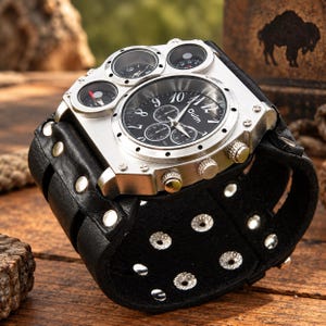 May include: A silver-toned wristwatch with a black dial featuring the brand name Oulm, three sub-dials, and two smaller circular dials at the top. The watch has a wide black leather cuff strap secured with silver-coloured metal rivets and studs.