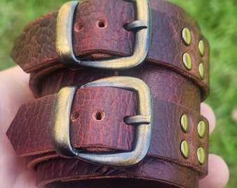 May include: A brown leather bracelet with two brass buckles and gold studs. The bracelet is made of two layers of leather.