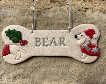 May include: Bone-shaped pet name sign with the name "BEAR" in silver lettering. The sign features a festive design with a dog wearing a Santa hat and coat, and a paw wearing a mitten. A holly sprig and berries are also included.