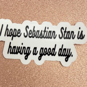 May include: A white sticker with the text "I hope Sebastian Stan is having a good day" in black cursive font. The sticker has a scalloped edge and is set against a rose gold background.