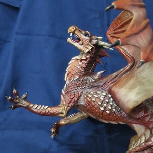 May include: A detailed figurine of a dragon in shades of red, brown, and tan. The dragon has textured scales, sharp claws, and large wings. Its mouth is open, revealing teeth. The dragon is posed dynamically, as if in mid-flight.