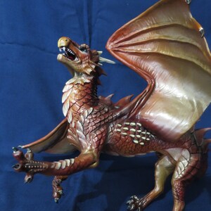 May include: A detailed figurine of a dragon with outstretched wings. The dragon is primarily reddish-brown with lighter accents on the wings and underbelly. Its mouth is open, revealing teeth, and it has sharp claws.