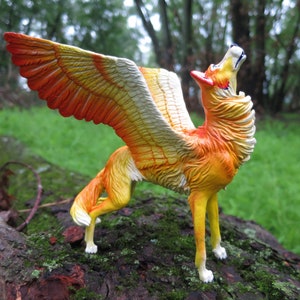 May include: A figurine of a winged wolf, with orange and yellow wings and fur. The wolf is posed howling upwards, with its head tilted back. The figurine is set on a mossy log against a blurred green background.