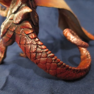 May include: A detailed sculpture of a dragon, featuring a textured, reddish-brown body and tail. The scales are meticulously crafted, with a curved tail extending from the body. The wings are visible in the background, with shades of orange and tan.