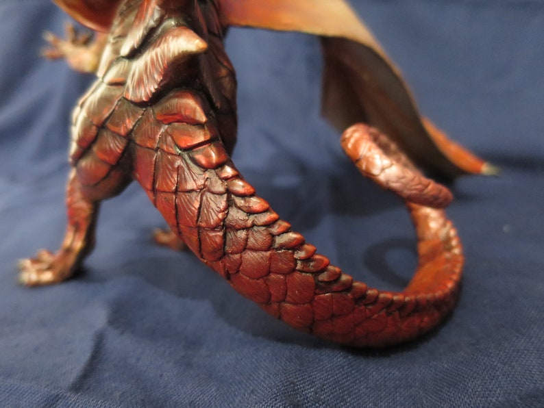 May include: A detailed sculpture of a dragon, featuring a textured, reddish-brown body and tail. The scales are meticulously crafted, with a curved tail extending from the body. The wings are visible in the background, with shades of orange and tan.