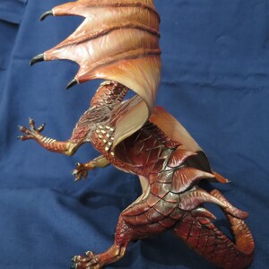 May include: A detailed figurine of a dragon in a dynamic pose. The dragon is primarily red with darker shading and gold accents. Its large wings are a mix of orange and brown. The dragon has sharp claws and a long tail, with a textured, scaled appearance.