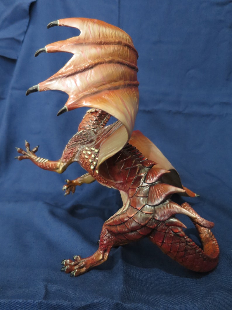 May include: A detailed figurine of a dragon in a dynamic pose. The dragon is primarily red with darker shading and gold accents. Its large wings are a mix of orange and brown. The dragon has sharp claws and a long tail, with a textured, scaled appearance.
