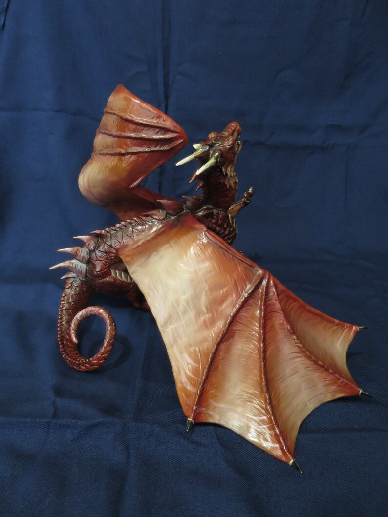 May include: A detailed, red and brown dragon figurine with outstretched wings. The dragon has a textured body, sharp claws, and a curled tail. The wings are a lighter shade of brown with detailed veining. The dragon is posed with its mouth open, as if roaring.