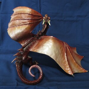 May include: A detailed sculpture of a dragon with outstretched wings and a curled tail. The dragon is primarily brown with lighter shades on the wings, and it appears to be breathing fire. The sculpture is set against a blue background.
