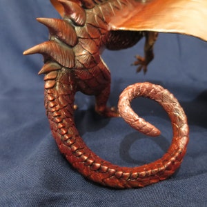 May include: A detailed sculpture of a dragon, featuring a curled tail and textured scales in shades of copper and bronze. The dragon's wing is visible in the background, with a blue background.