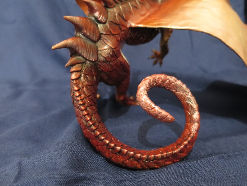May include: A detailed sculpture of a dragon, featuring a curled tail and textured scales in shades of copper and bronze. The dragon's wing is visible in the background, with a blue background.