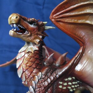 May include: A detailed, red and brown dragon figurine with open mouth, sharp teeth, and detailed scales. The dragon has large wings and is set against a blue background. The figurine is a fantasy-themed collectible.