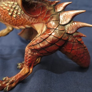 May include: A detailed sculpture of a red and brown dragon with textured scales and sharp spines. The dragon is in a dynamic pose, with its claws extended and wings partially visible. The sculpture has a detailed, realistic appearance.