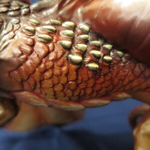 May include: Close-up of a detailed sculpture featuring textured, reddish-brown scales with gold-colored accents. The sculpture has a reptilian or mythical creature theme, with visible ridges and a smooth, polished surface.