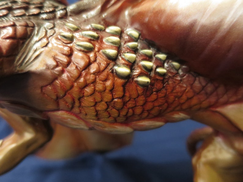 May include: Close-up of a detailed sculpture featuring textured, reddish-brown scales with gold-colored accents. The sculpture has a reptilian or mythical creature theme, with visible ridges and a smooth, polished surface.