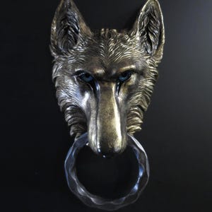 May include: A bronze-coloured wolf head door knocker with blue eyes and a textured finish. The knocker has a ring-shaped handle. The wolf's head is mounted on a black surface.
