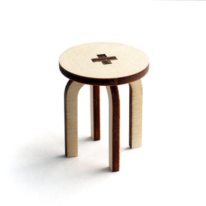 May include: A small wooden stool with a round top and three legs. The top has a brown plus sign design.