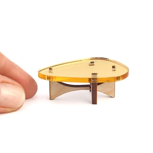 May include: A miniature coffee table featuring a translucent amber-colored acrylic top with a teardrop shape. The base is constructed from dark brown wood with a curved, intersecting support structure that holds the tabletop securely in place.
