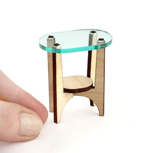 May include: Miniature wooden side table with a clear, oval-shaped glass top. The table has a light wood finish and a small, round shelf. The table is approximately 2 inches tall.