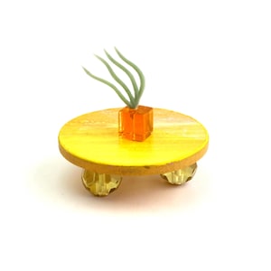 May include: A small, round, bright yellow platform stand supported by three faceted, translucent amber-colored bead feet. Centered on the yellow surface is a small, translucent orange cube holding a single, pale green, multi-stemmed air plant.