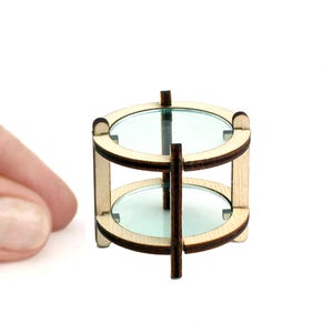 May include: Miniature two-tiered table with a light wood frame and clear glass shelves. The table has a round shape and is designed for a dollhouse or other small-scale setting. The table is a light brown color.