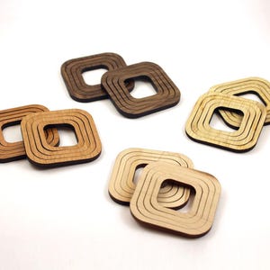 May include: A collection of wooden square coasters in various shades of brown and tan. Each coaster features a square cutout in the center and concentric square grooves. The coasters are arranged on a white surface, showcasing their natural wood grain.