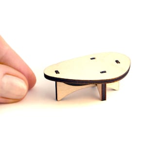Könnte beinhalten: Ein Miniatur-Couchtisch aus hellem Holz mit dunkelbraunen Kanten. Der Tisch hat eine ovale Oberseite mit vier kleinen quadratischen Aussparungen und drei Beinen. Der Tisch ist etwa 5 cm lang.