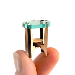 Puede incluir: Una mesa auxiliar en miniatura con una superficie circular de acrílico verde claro translúcido, sostenida por cuatro patas de madera marrón oscuro y un pequeño estante redondo de madera situado entre las patas, creando un mueble compacto de varios niveles.