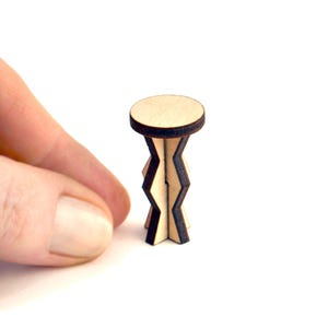 May include: A miniature wooden side table with a round top and a zigzag-patterned base. The table is made of light-colored wood with dark brown edges. The table is held by a hand against a white background.