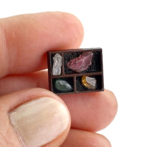 May include: A miniature wooden display case with four compartments, each holding a different gemstone. The gemstones are various colors, including clear, pink, green, and yellow. The case is held in a person's hand.