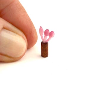 May include: A tiny wooden vase with pink flowers. The vase is cylindrical and has a dark brown finish. The flowers are a translucent pink colour and have a delicate appearance. The overall aesthetic is delicate and charming.