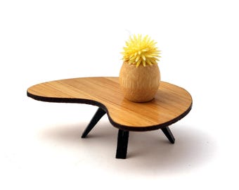 May include: A miniature mid-century modern style coffee table with a light wood grain finish. The table has a kidney bean shape with black legs. A small wooden vase with a yellow flower sits on top.