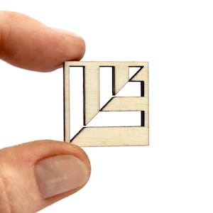 Puede incluir: Una pequeña pieza de rompecabezas cuadrada de madera con un diseño geométrico. La pieza se sostiene entre los dedos sobre un fondo blanco. El rompecabezas presenta formas y líneas recortadas.