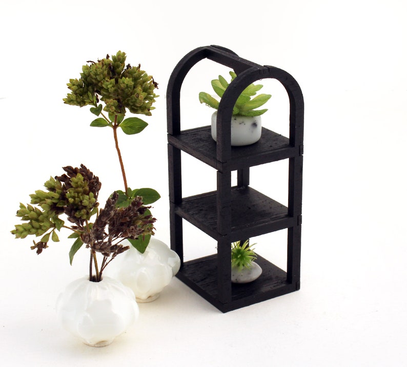May include: A black wooden shelf unit with three shelves, each holding a small potted plant. The plants are green and the pots are white. The shelf unit is on a white background.