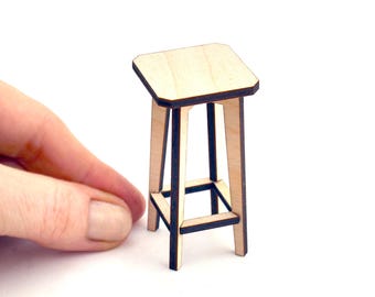 May include: A miniature wooden stool with a light-colored seat and legs. The stool has a geometric design with a dark outline. The stool is approximately 3 inches tall.