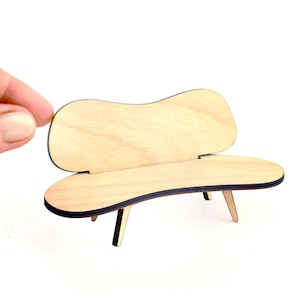 May include: A miniature wooden bench with a curved back and seat, supported by three angled legs. The bench is made of light-colored wood with dark edges. The design is mid-century modern, perfect for a dollhouse or miniature scene.
