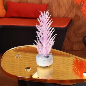 May include: Miniature interior scene featuring a gold-colored, oval coffee table with a small, light purple plant in a gray and white pot. The table reflects the plant and has small, square decorative accents.