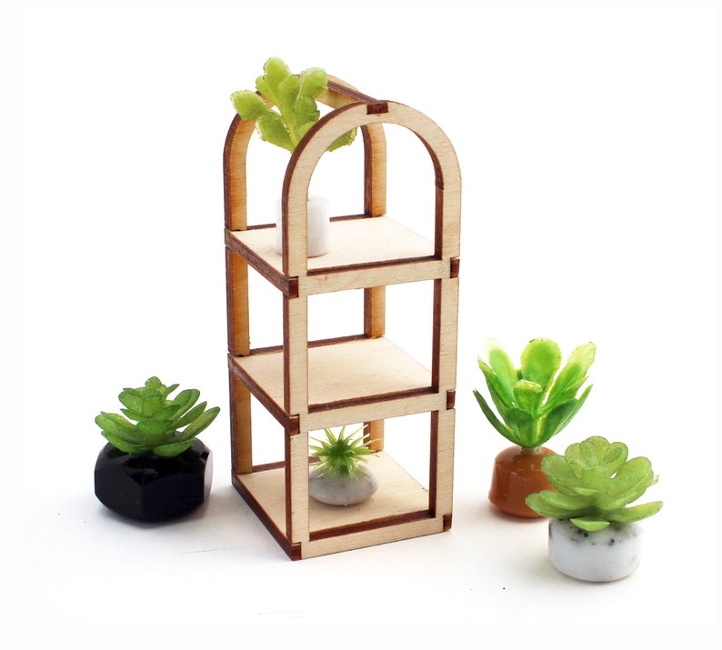 May include: A wooden shelf unit with three shelves and an arched top. The shelf unit is empty except for a small white pot on the top shelf. There are three small potted plants in front of the shelf unit.