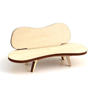 May include: A miniature wooden bench with a curved back and seat. The bench has three angled legs and a light brown finish with darker brown edges. The design is simple and modern, suitable for dollhouses or craft projects.
