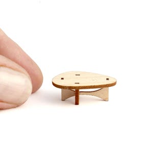 Könnte beinhalten: Ein Miniatur-Couchtisch aus hellem Holz. Der Tisch, etwa 4 cm im Durchmesser, hat eine abgerundete Form mit vier kleinen quadratischen Aussparungen auf der Oberseite. Er hat ein zentrales Bein und gebogene Stützen, für ein Puppenhaus.