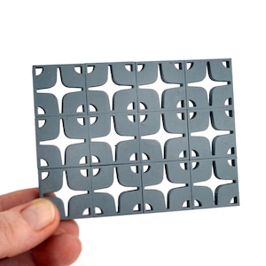 May include: A gray, geometric decorative panel with a repeating pattern of square shapes with rounded corners and circular cutouts. The panel is held in a hand against a white background. The design is reminiscent of mid-century modern aesthetics.